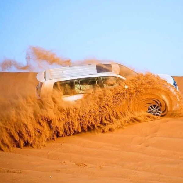 White SUV driving across vast sandy dunes, kicking up dust