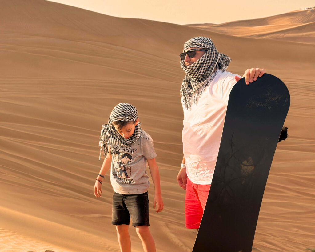 A man and a boy prepare for sandboarding in a desert landscape