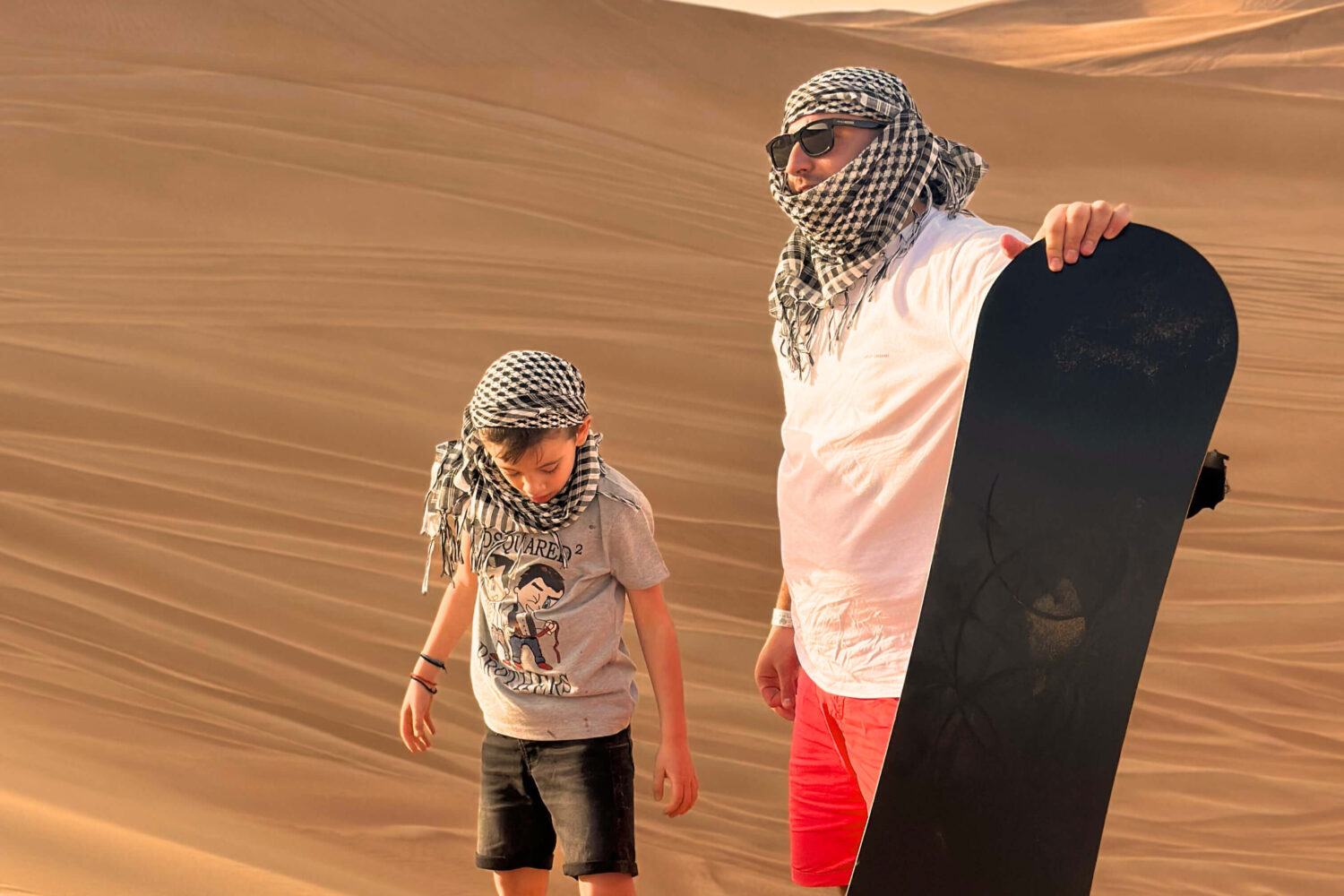 A man and a boy prepare for sandboarding in a desert landscape