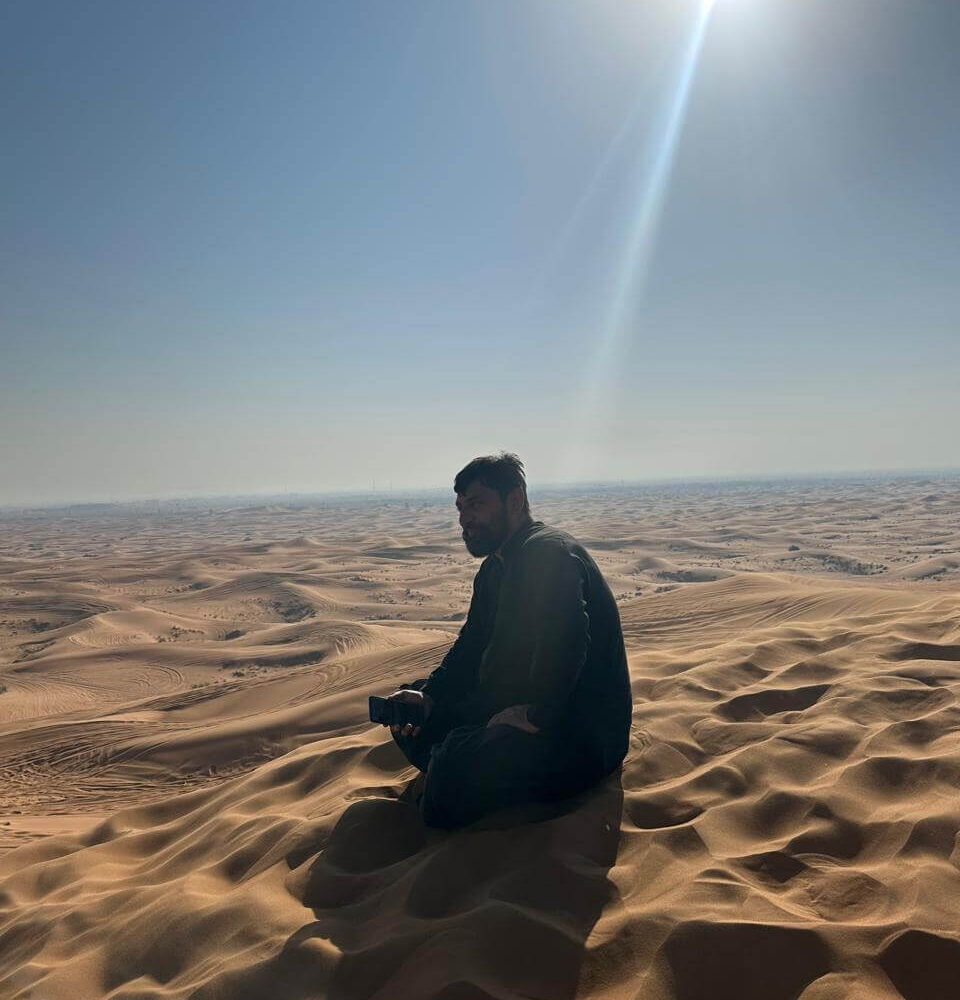 Man sits in the sunlit desert, surrounded by vast sand dunes